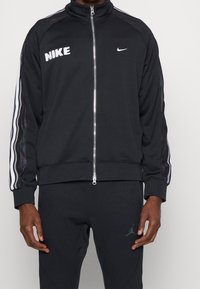 Chaqueta con cremallera negra de Nike con logo blanco y rayas laterales. Tejido suave, puños y dobladillo acanalados, cuenta con un detalle reflectante en las mangas.