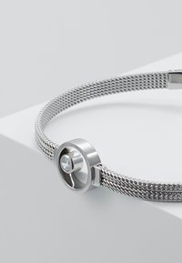 Silbernes Mesh-Armband mit einem runden Metallelement. Besitzt einen zentralen klaren Edelstein und eine glatte, polierte Oberfläche. Verstellbarer Verschluss.