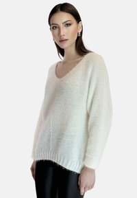 Witte gebreide trui met een losse pasvorm, V-hals en geribbelde zoom. Zachte textuur en wijdere mouwen. Gepaard met zwarte leggings.