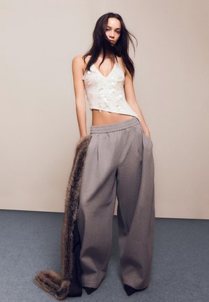 Mladá žena, ktorá má na sebe biele flitrové halter top, široké sivé nohavice a drží dlhý kožuch na béžovom pozadí.