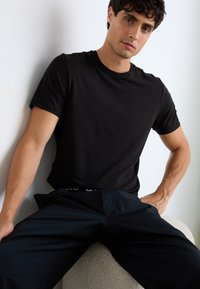 T-shirt preta de gola redonda, feita de tecido suave, combinada com calças azul-marinho escuras com um cós elástico com marca. Modelo sentado numa superfície clara.