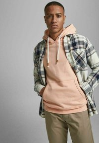 Karierte Flanellhemd in Blau, Weiß und Grün über einem Pfirsich-Hoodie mit Kordelzügen; beigen Hosen; männliches Model mit kurzem Haar.