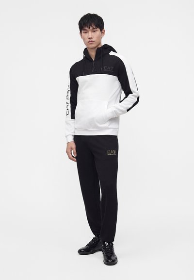 Мъж, стоящ, носещ черно-бяла качулка EA7 Emporio Armani и черен спортен панталон с черни маратонки на бял фон.