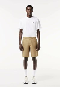 Pantaloncini kaki con chiusura a bottone e passanti per cintura, abbinati a una maglietta bianca semplice con un piccolo logo verde. Scarpe da ginnastica bianche in mostra.