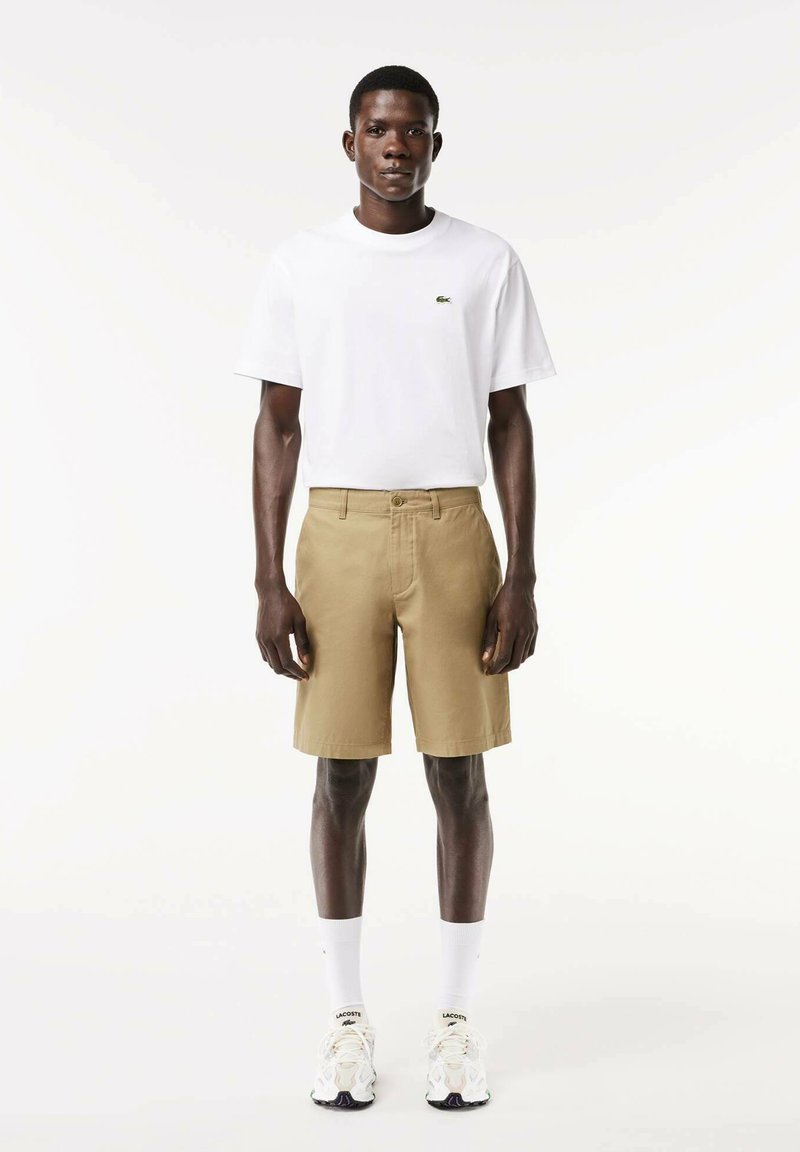 Pantaloncini kaki con chiusura a bottone e passanti per cintura, abbinati a una maglietta bianca semplice con un piccolo logo verde. Scarpe da ginnastica bianche in mostra.