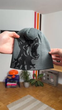 Pelēks adīts cepure ar melnu attēlu, kurā ir redzams aurojošs vilks. Mīksta tekstūra un cieši adīts raksts, kas paredzēts siltumam un ērtai piekļaušanai.