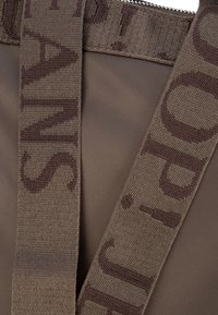 Hnedá tkaninová taška so štruktúrovaným dizajnom, ktorá má textúrovaný hnedý popruh s potlačou. Detail zipsu je viditeľný na vrchu.