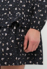 Μαύρο φόρεμα με ανθικό σχέδιο και βολάν στη μέση, με μικρά μπλε και μπεζ λουλούδια και ελαστικές μανσέτες, κατασκευασμένο από ελαφρύ ύφασμα.