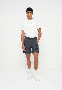 Outfit da uomo composto da una t-shirt bianca con logo multicolore, pantaloni corti grigi scuri con tasche laterali e sneakers bianche in tessuto texturizzato.