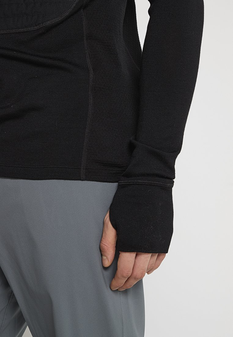 Schwarzes Langarmoberteil mit Daumenlöchern, hergestellt aus einem glatten, dehnbaren Material. Genähte Details sind an den Nähten sichtbar. Getragen mit grauen Hosen.
