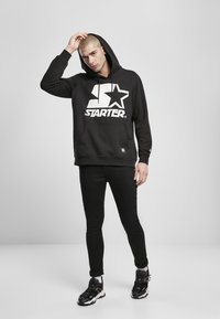 Svart hettegenser med stort hvitt "STARTER"-logo og stjernesymbol, kengurulomme, snordrag, brukt med svarte slim fit jeans og sneakers.
