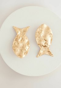 Piezas decorativas en forma de pez de tono dorado con una textura martillada, colocadas sobre una superficie blanca y redonda. Cada pez tiene diseños de cola distintos.