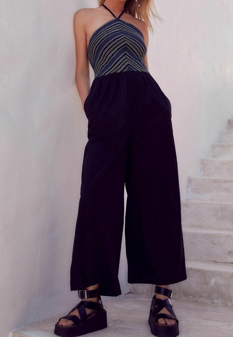 Femme portant une combinaison sans manches avec un haut à motifs et un pantalon large noir, debout près d'escaliers en pierre blanche, les mains dans les poches.