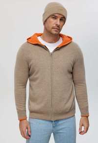 Beige zip-up hoodie with an orange lining, ribbed cuffs, and a soft texture, paired with a matching beige knit beanie.