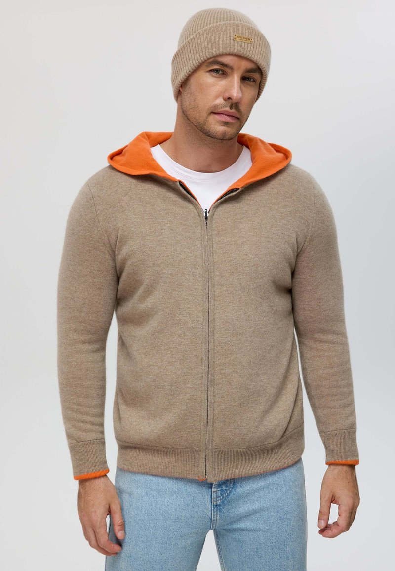Beige zip-up hoodie with an orange lining, ribbed cuffs, and a soft texture, paired with a matching beige knit beanie.