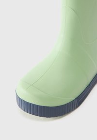 Bota de goma verde con superficie lisa, punta redondeada y suela texturizada de color azul oscuro. Presenta un diseño sin costuras y material suave y flexible.