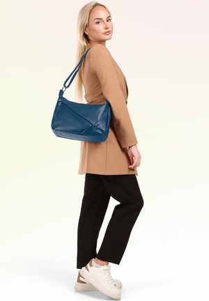 Femme en blazer beige et pantalon noir porte un sac à bandoulière en cuir bleu, portant des baskets blanches, regarde par-dessus son épaule.