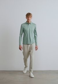 Világoszöld hosszú ujjú pólóing krémszínű, karcsúsított nadrággal és fehér tornacipővel párosítva. Egyszerű dizájn, puha textúra és finom márkajelzés.