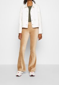 Weiße Jeansjacke, grünes geripptes Oberteil, beige Cord-Flared-Hose und weiße Sneaker mit roten Akzenten. Einfache Knopfdetails an der Jacke.