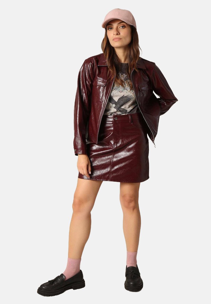 Ensemble en simili cuir bordeaux comprenant une veste et une jupe, avec un T-shirt graphique, une casquette rose et des chaussures à plateforme noires avec des chaussettes roses.