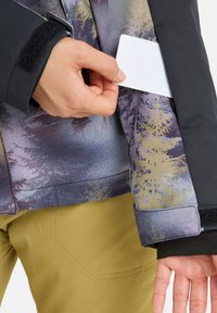 Black jacket with a colorful, patterned texture showing trees, featuring a Velcro cuff and a hand inserting a card into a side pocket.