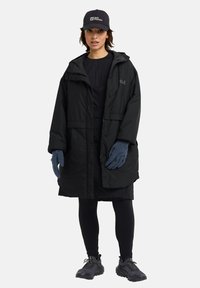 Parka noire rembourrée avec capuche, gants foncés et leggings noirs. Le modèle porte une casquette sombre avec un logo. Des chaussures robustes complètent la tenue.