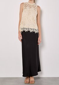 Haut sans manches en dentelle crème avec motifs floraux, associé à une longue jupe noire et des sandales à lanières, présentant une texture lisse et un design fluide.