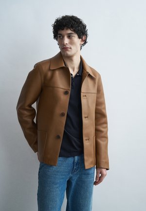 Hombre con chaqueta de cuero marrón sobre una camiseta negra y jeans azules, de pie contra un fondo claro y liso, mirando hacia la izquierda.