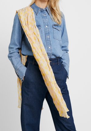 Personne blonde portant une chemise en denim bleu rentrée dans un jean foncé, les mains dans les poches, et une écharpe rose pâle avec des motifs jaunes drapée sur l'épaule.