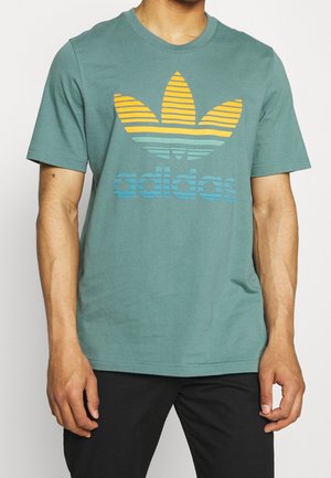 Zielona bawełniana koszulka z krótkim rękawem, z okrągłym dekoltem i paskowanym logo adidas w pomarańczowym i niebieskim kolorze.