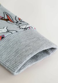 Jasnoszara mankiety bluzy z ribbowym splotem i nadrukowanym rysunkowym wzorem przedstawiającym postać. Miękka tkanina, złożona na krawędzi.