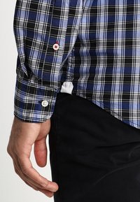 Mano maschile appoggiata accanto a pantaloni neri, indossando una camicia a scacchi blu e nera con bottoni bianchi e rossi.