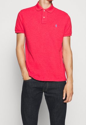 Mann trägt ein lebhaftes rotes Polohemd mit kurzen Ärmeln und einem kleinen blauen gestickten Logo auf der linken Brust sowie dunkle Jeans.