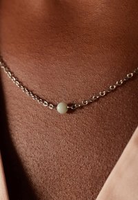 Zilveren ketting met een eenvoudig ontwerp, uitgerust met een kleine witte kraal als accent, rustend tegen een getextureerde, bruine achtergrond.