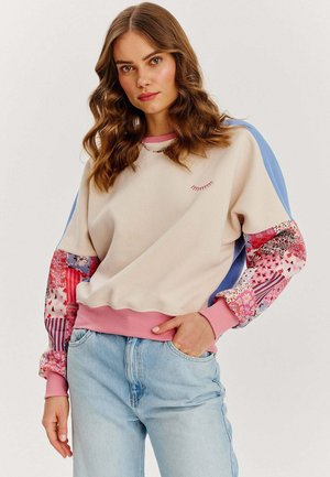 Bluza z kremowym korpusem, różowymi mankietami i kolorowymi wzorzystymi rękawami. Zawiera małe dekoracyjne przeszycie z przodu.
