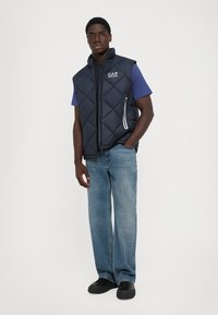 Gilet matelassé marine avec poches zippées, porté sur un t-shirt bleu, associé à un jean bleu clair à coupe ample et des chaussures noires.
