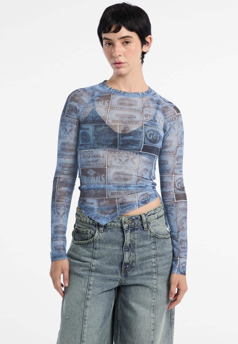 Langzaam mouwloze doorschijnende top met een blauwe grafische print van verschillende labels en texturen, gecombineerd met losse, lichtgekleurde denim broek.