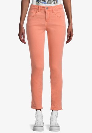 Jeans de color durazno con un diseño ajustado, cinco bolsillos y un dobladillo ligeramente deshilachado. Combinados con zapatillas blancas.