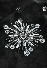 Sort stof med indviklede sølvfarvede paletter arrangeret i et solstrålemønster, med varierende størrelser af perler og blomsterlignende designs indlejret.