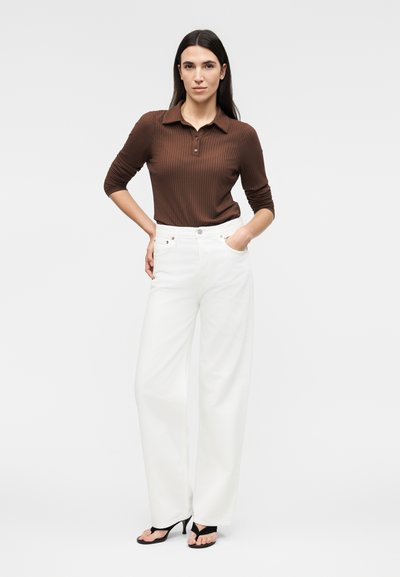 Γυναίκα που φοράει καφέ ριμπ ζιβάγκο με μακριά μανίκια και λευκό παντελόνι με φαρδιά πόδια, μαύρα σανδάλια με τακούνι, στέκεται με το ένα χέρι στην τσέπη.