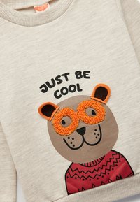 Jasno-beżowy sweatshirt z grafiką misia w pomarańczowych okularach, czarnymi akcentami i czerwonym wzorem dzianiny. Napis brzmi "PO PROSTU BĄDŹ FAJNI".