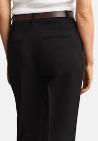 Persoană care poartă pantaloni negri croiți, cu buzunare tăiate în spate, curea maro și top alb fără mâneci, cu model tip rib, văzută din spate de la talie în jos.