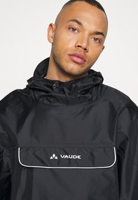 Črna vodoodporna jakna s kapuco in belimi detajli, opremljena s sprednjim žepom in prominentnim logotipom "VAUDE". Gladka tekstura.