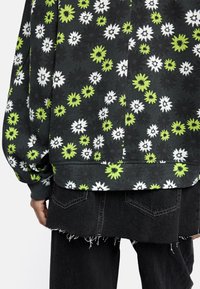 Bluza z czarnym tłem, zdobiona kwiatowym wzorem w białym i zielonym kolorze, z ściągaczami przy rękawach i prostym dołem, zestawiona z postrzępionymi czarnymi dżinsami.