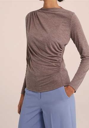 Femme portant un haut marron ajusté à manches longues avec fronces latérales et pantalon bleu clair taille haute, main dans la poche.
