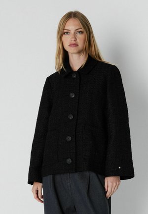 Une femme porte une veste noire texturée, boutonnée sur le devant et dotée de manches amples, ainsi qu’un pantalon foncé, sur un fond gris uni.