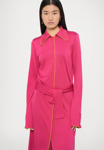 Fuksjowa sukienka z długim rękawem, z ostrokątnym kołnierzem, żółtym przeszyciem, guzikami z przodu oraz paskiem wiązanym w talii. Gładka tkanina o luźnym kroju.