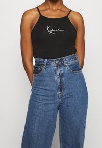 Canotta nera con spalline sottili che presenta un testo bianco scritto a mano. Abbinata a jeans in denim blu a vita alta.