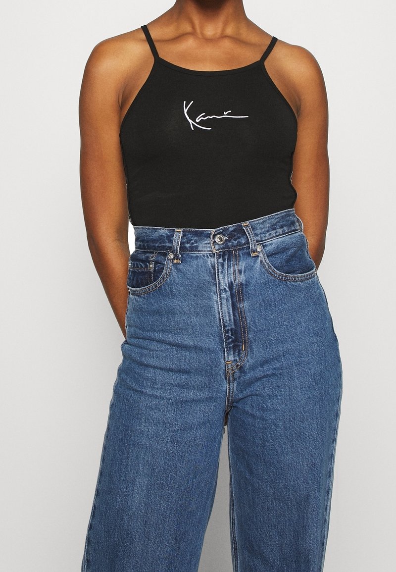 Canotta nera con spalline sottili che presenta un testo bianco scritto a mano. Abbinata a jeans in denim blu a vita alta.