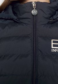 Laivastonsininen toppatakki, jossa on korkea kaulus, kontrastivetoketju ja rintamerkki. Takissa on pehmeä, teksturoitu kangas.